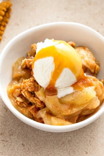 A bowl of apple crisp topped with a scoop of vanilla ice cream and drizzled with caramel sauce, served on a beige surface.