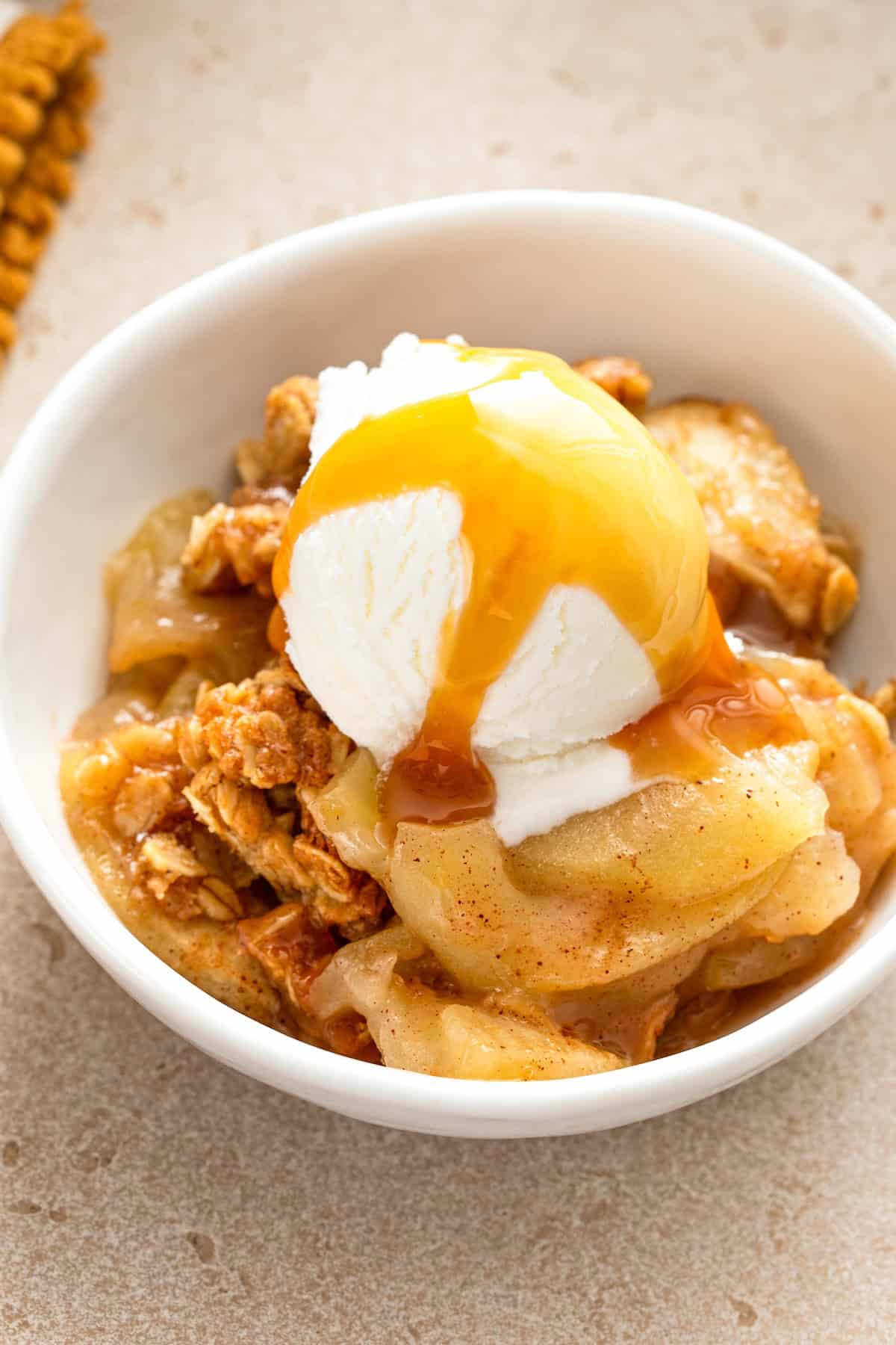 A bowl of apple crisp topped with a scoop of vanilla ice cream and drizzled with caramel sauce, served on a beige surface.