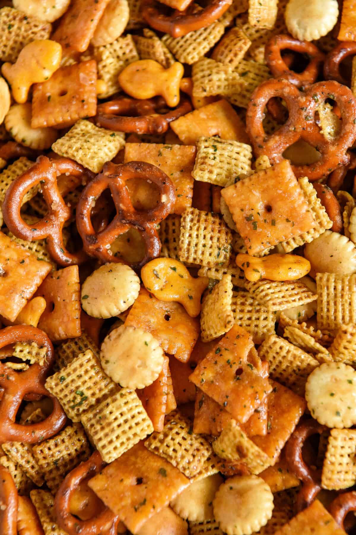 A close-up of cheddar chex mix including pretzels, cheese crackers, oyster crackers, fish-shaped crackers, and seasoned cereal squares.