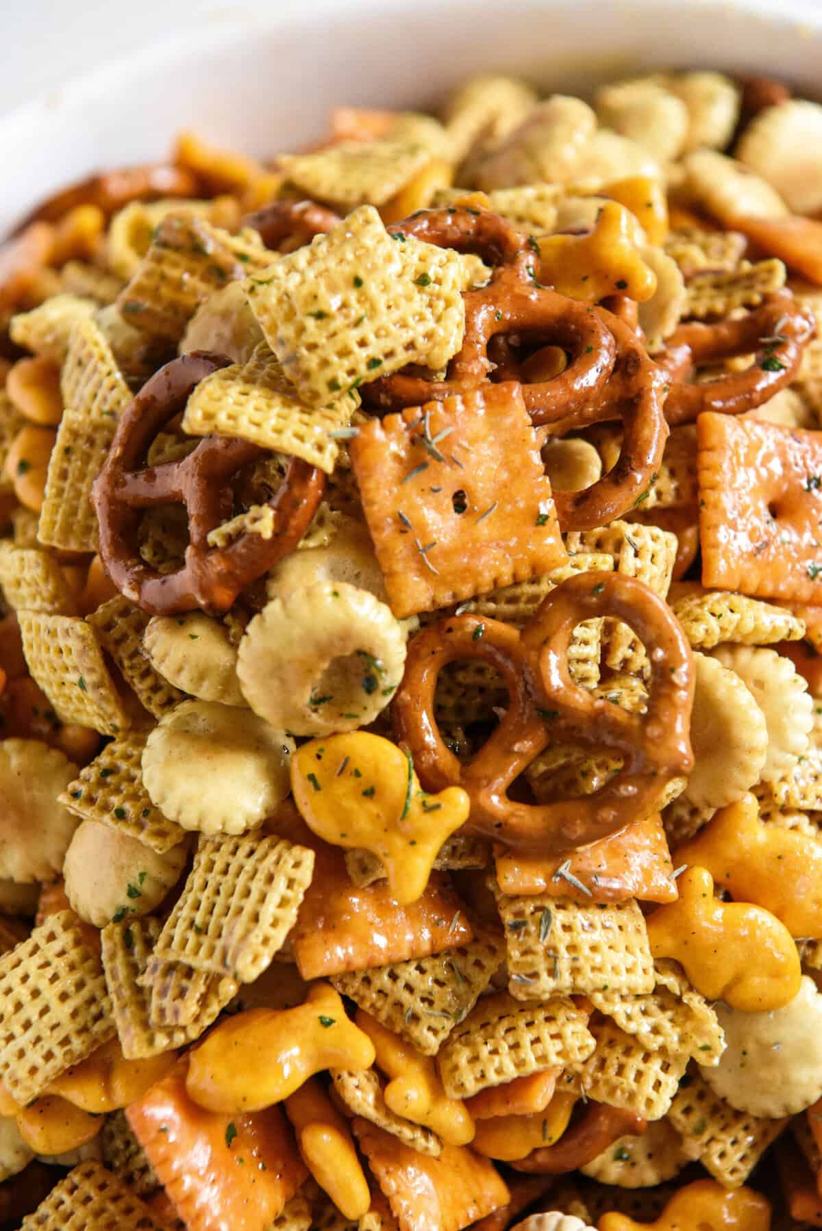 A close-up of a homemade chex mix recipe being seasoned and mixed together in a bowl with pretzels, Chex cereal, cheese crackers, oyster crackers, and goldfish crackers.