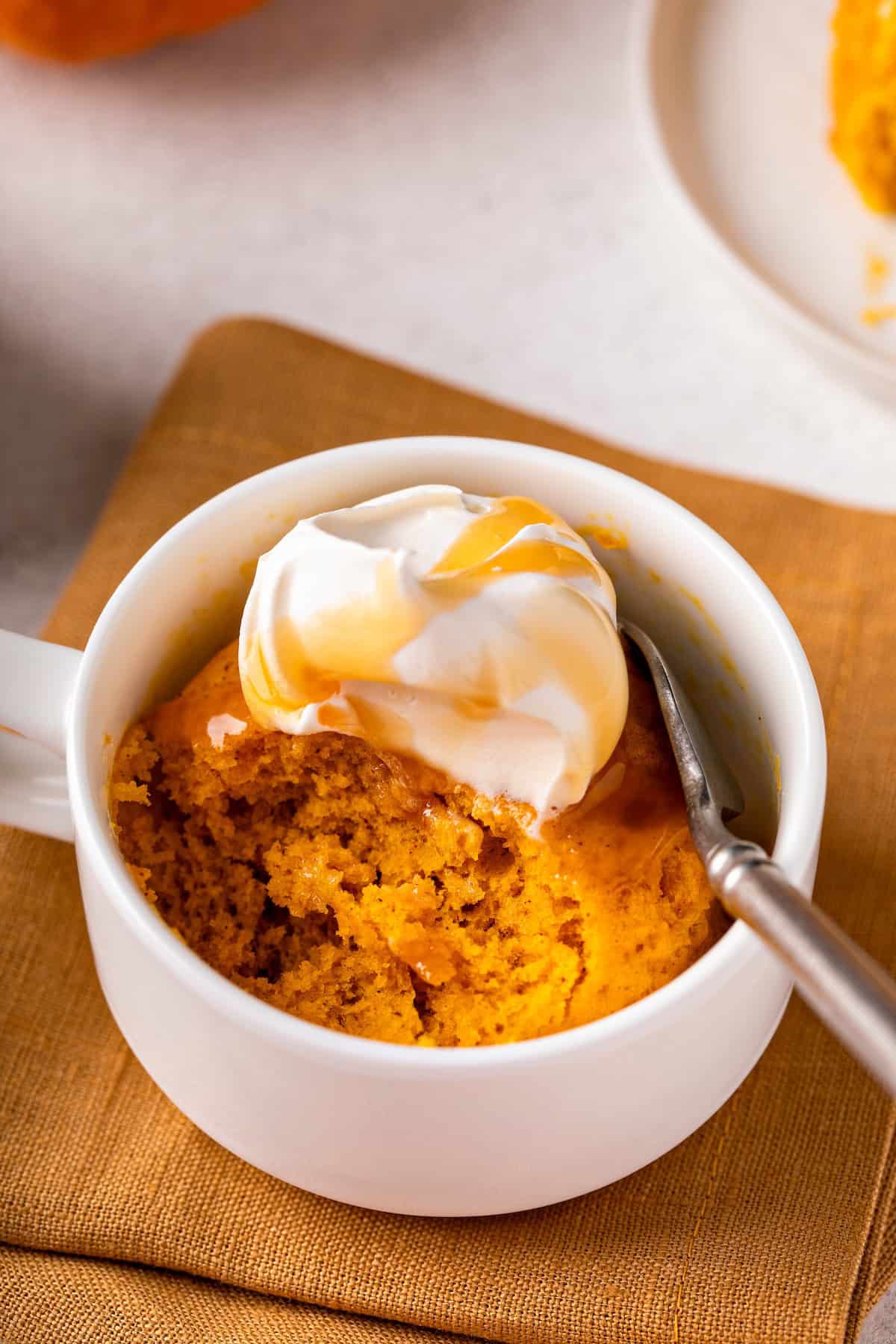 A white mug filled with pumpkin mug cake, topped with whipped cream and caramel sauce, sits on a brown napkin with a spoon inside the mug.