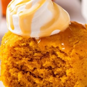 A moist pumpkin mug cake topped with a dollop of whipped cream and drizzled with caramel sauce, served on a white plate.