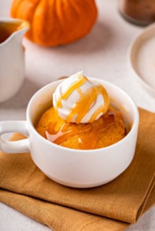 A white mug filled with pumpkin mug cake topped with whipped cream and caramel sauce, placed on an orange cloth with a small pumpkin and dish in the background.