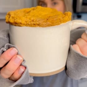 A person in a gray sweater holds a large beige mug filled with a fluffy baked treat, possibly pumpkin mug cake, with the cake risen above the rim.