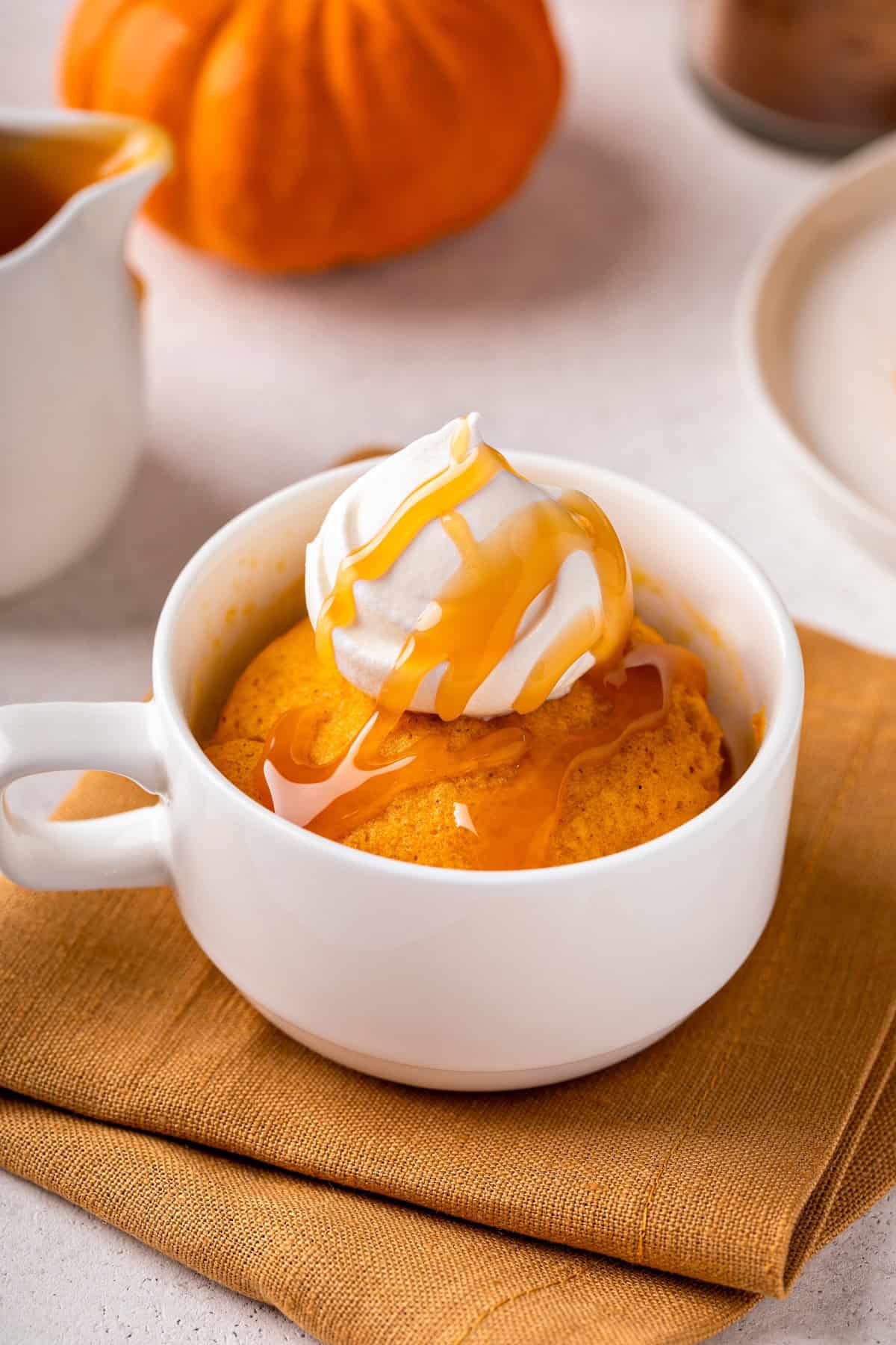 Fluffy, moist, and perfectly spiced, this Pumpkin Mug Cake is made with simple pantry staples (no eggs!) and ready in just 5 minutes! A white mug filled with pumpkin mug cake topped with whipped cream and caramel sauce, placed on an orange cloth with a small pumpkin and dish in the background.
