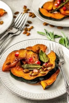 Slices of roasted acorn squash topped with pomegranate seeds, toasted pumpkin seeds, and fresh sage leaves are served on a white plate with a fork next to the dish.