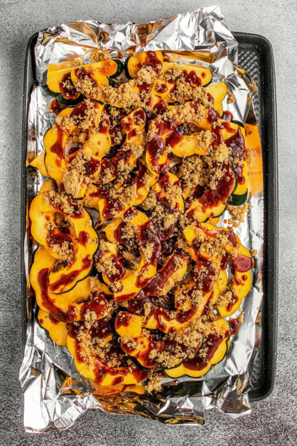 Sliced acorn squash on a foil-lined baking sheet, topped with brown sugar and drizzled with a spiced sauce mixture, ready to be baked.