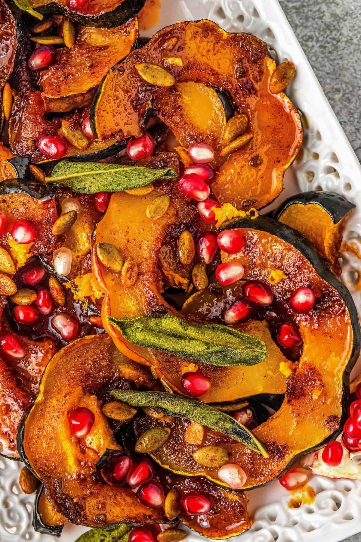 Sliced roasted acorn squash garnished with crispy sage leaves, pumpkin seeds, and bright pomegranate arils, served on a decorative white platter.
