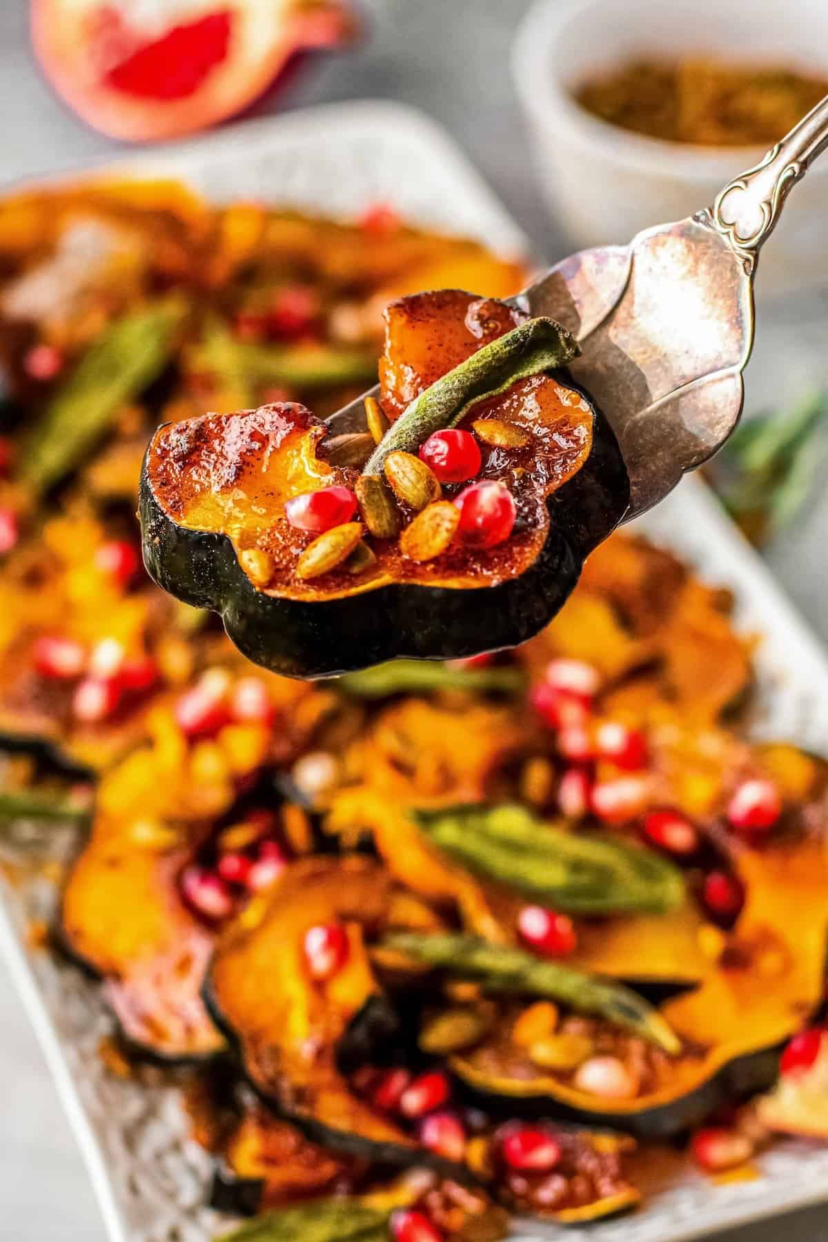 A close-up of roasted squash garnished with pomegranate seeds, pumpkin seeds, and fresh herbs on a silver serving utensil, with more squash on a platter in the background.
