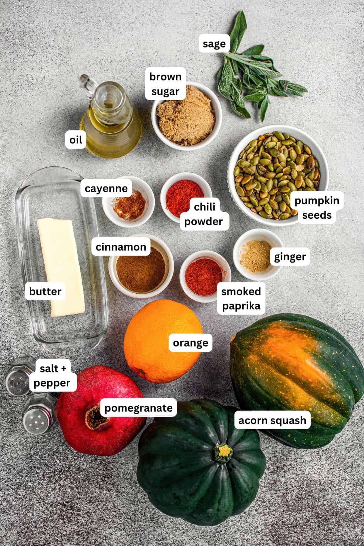 Overhead view of labeled ingredients for roasted acorn squash recipe on a gray surface: sage, oil, brown sugar, pumpkin seeds, ginger, smoked paprika, chili powder, cayenne, cinnamon, butter, salt, pepper, orange, pomegranate, and two acorn squash.