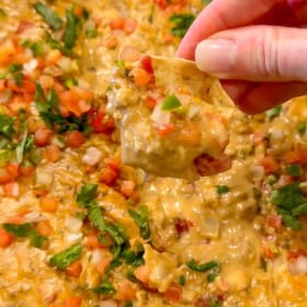 A hand holds a tortilla chip loaded with cheesy queso dip, ground beef, diced tomatoes, onions, and chopped cilantro over a dish of the same dip.