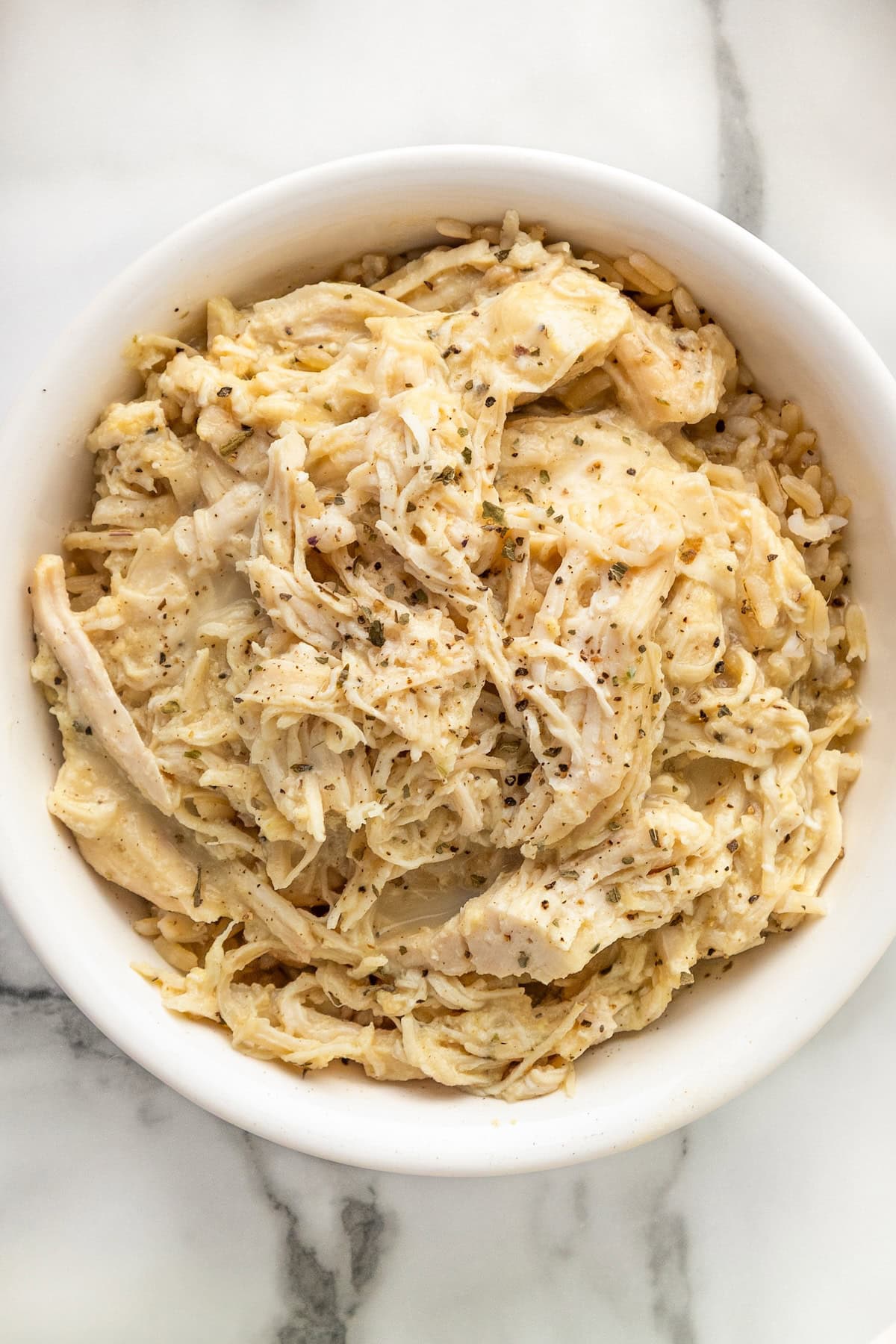 Skip the canned soup! This easy Crockpot Chicken and Gravy recipe is made from scratch - just real ingredients, creamy gravy, and big flavor! A white bowl filled with creamy shredded crockpot chicken and gravy seasoned with herbs and pepper, served over a bed of rice, on a marble surface.