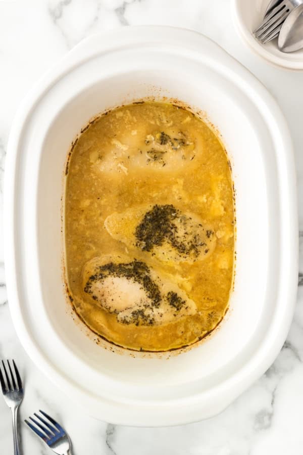 Three seasoned chicken breasts cooked in a creamy gravy inside a white slow cooker, viewed from above, with two forks on the side.