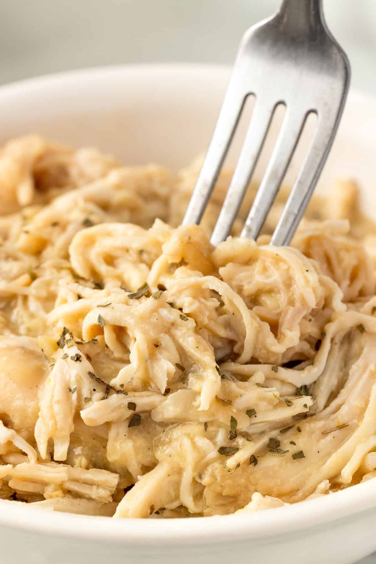 A close-up of a fork holding a portion of crockpot chicken and gravy, garnished with herbs, served in a white bowl.