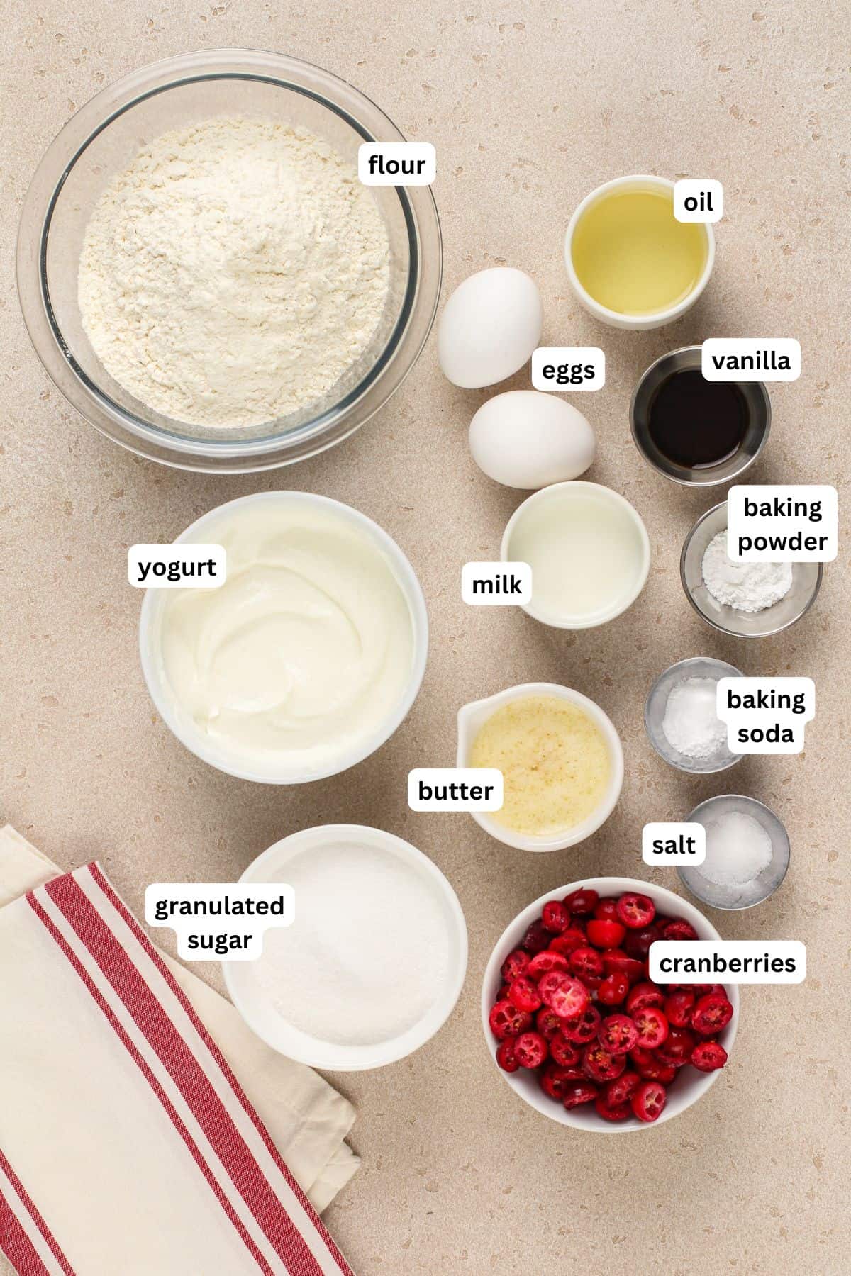 Top-down view of cranberry muffin recipe ingredients arranged on a counter, labeled: flour, eggs, oil, vanilla, yogurt, milk, baking powder, baking soda, butter, salt, granulated sugar, and cranberries. A striped towel is on the side.