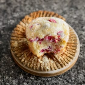 A cranberry muffin with a bite taken out of it sitting on a muffin wrapper on a speckled gray surface.