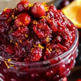 A close-up of a glass bowl filled with chunky cranberry sauce, topped with whole cranberries and orange zest. An orange wedge is visible in the blurred background.