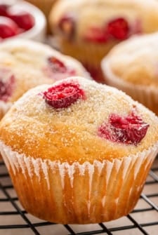 A close-up of a golden cranberry muffin with a sprinkle of sugar on top, resting on a cooling rack, with more muffins and a bowl of cranberries in the background.