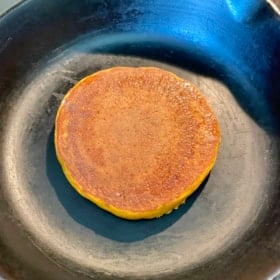 A thick, round pumpkin pancake with a golden-brown surface cooks in a black cast iron skillet.