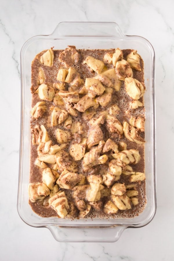 Make easy with just 6-ingredients, every bite of this Cinnamon Roll Casserole tastes like the gooey and warm center of a cinnamon roll! A glass casserole dish filled with chopped cinnamon rolls and an egg, cinnamon, and milk mixture, ready to be baked.