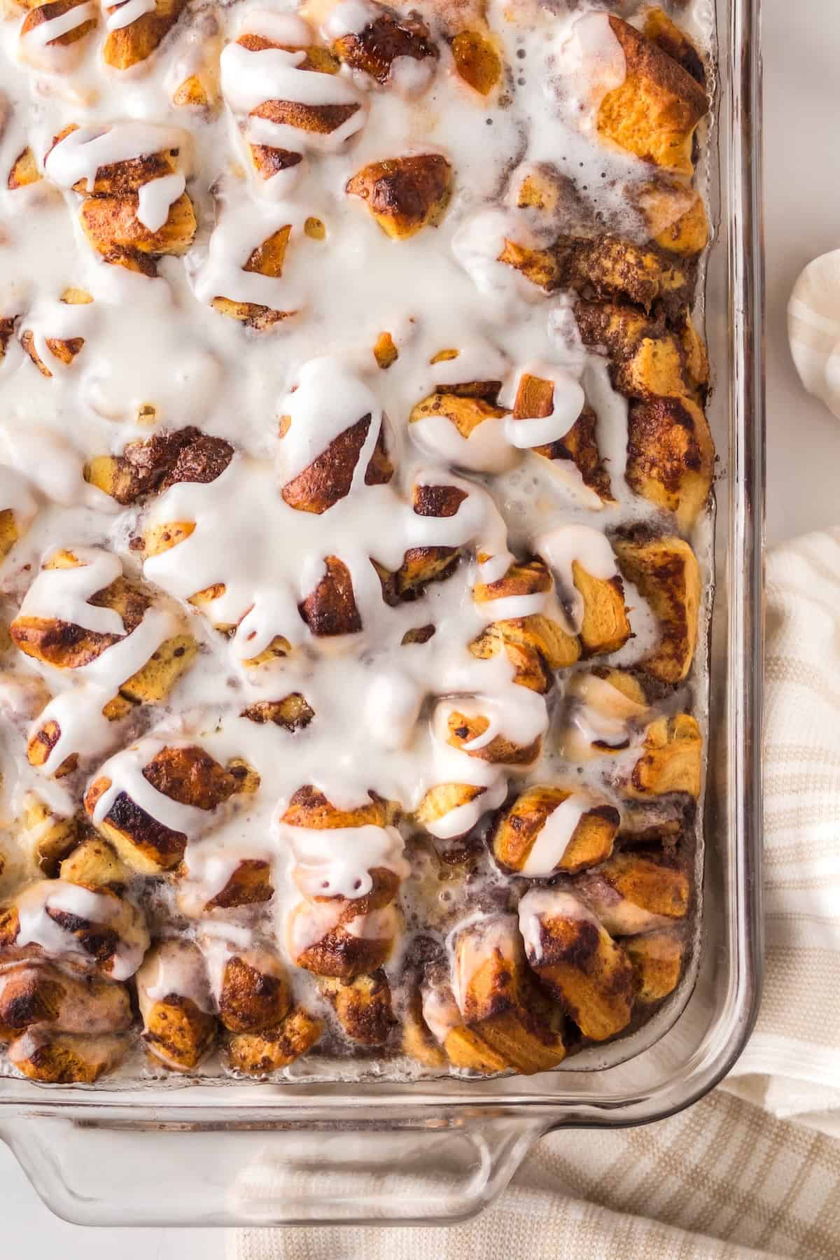 Make easy with just 6-ingredients, every bite of this Cinnamon Roll Casserole tastes like the gooey and warm center of a cinnamon roll! A close-up of a glass baking dish filled with cinnamon roll casserole drizzled with white icing.