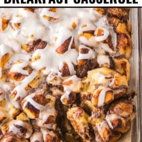 A close-up of cinnamon roll breakfast casserole in a glass baking dish, topped with white icing, with a wooden spoon scooping out a portion.