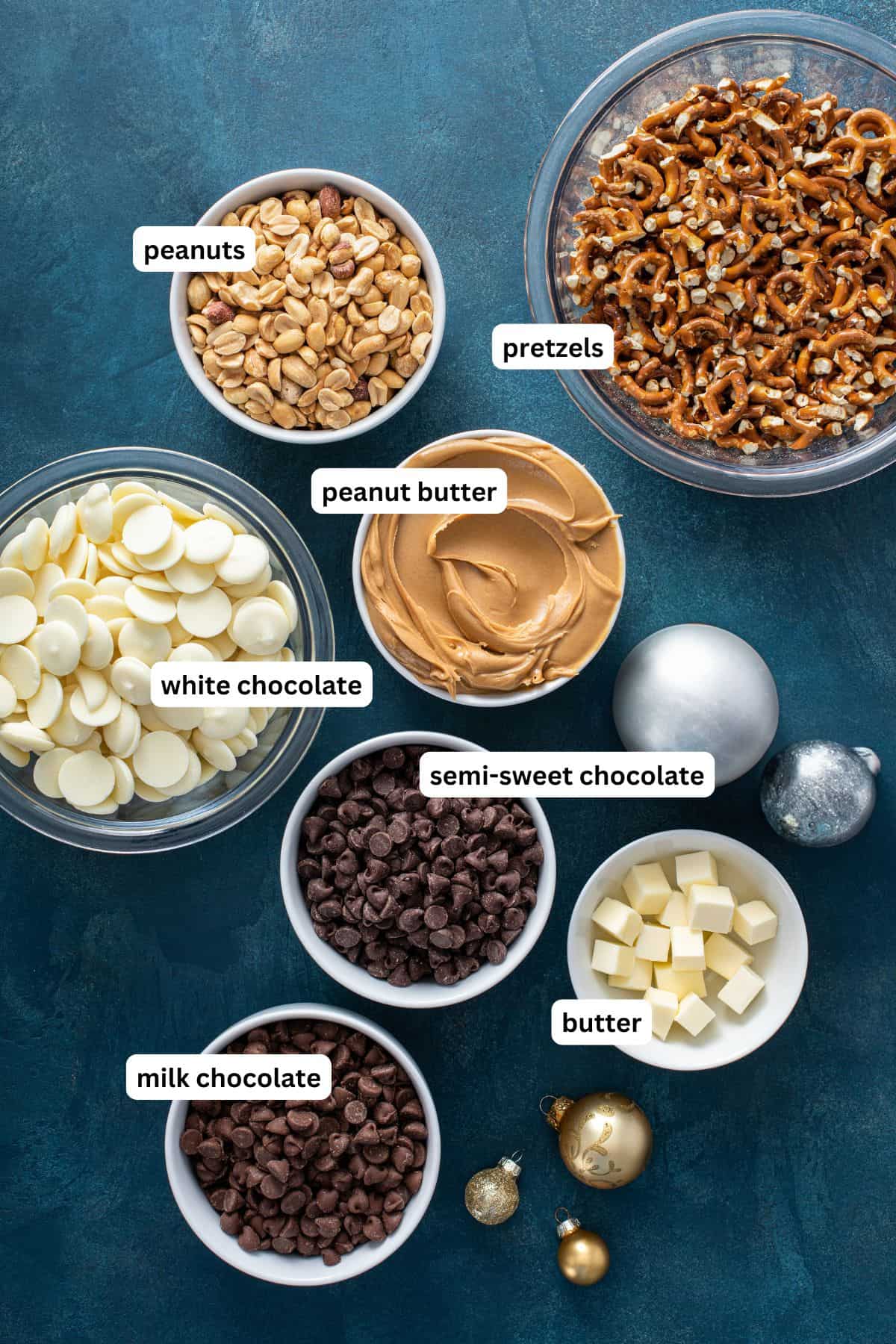 Simply toss all of the ingredients into the crockpot, stir a couple of times, scoop, and bam! Homemade Peanut Butter Pretzel Crockpot Candy! Bowls containing labeled ingredients for this crockpot candy recipe: peanuts, pretzels, white chocolate, peanut butter, semi-sweet chocolate chips, milk chocolate chips, and butter.