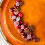 A caramel colored eggnog flan topped with sugared cranberries and star anise, arranged decoratively on one side, served on a glass platter.
