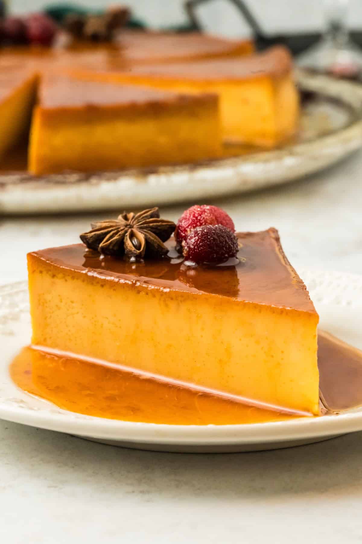 This festive Eggnog Flan is smooth, rich, and creamy, with plenty of nutmeg and rum for that classic spiced holiday flavor. A slice of eggnog flan on a white plate, topped with a star anise and a sugared berry, with the rest of the flan in the background.