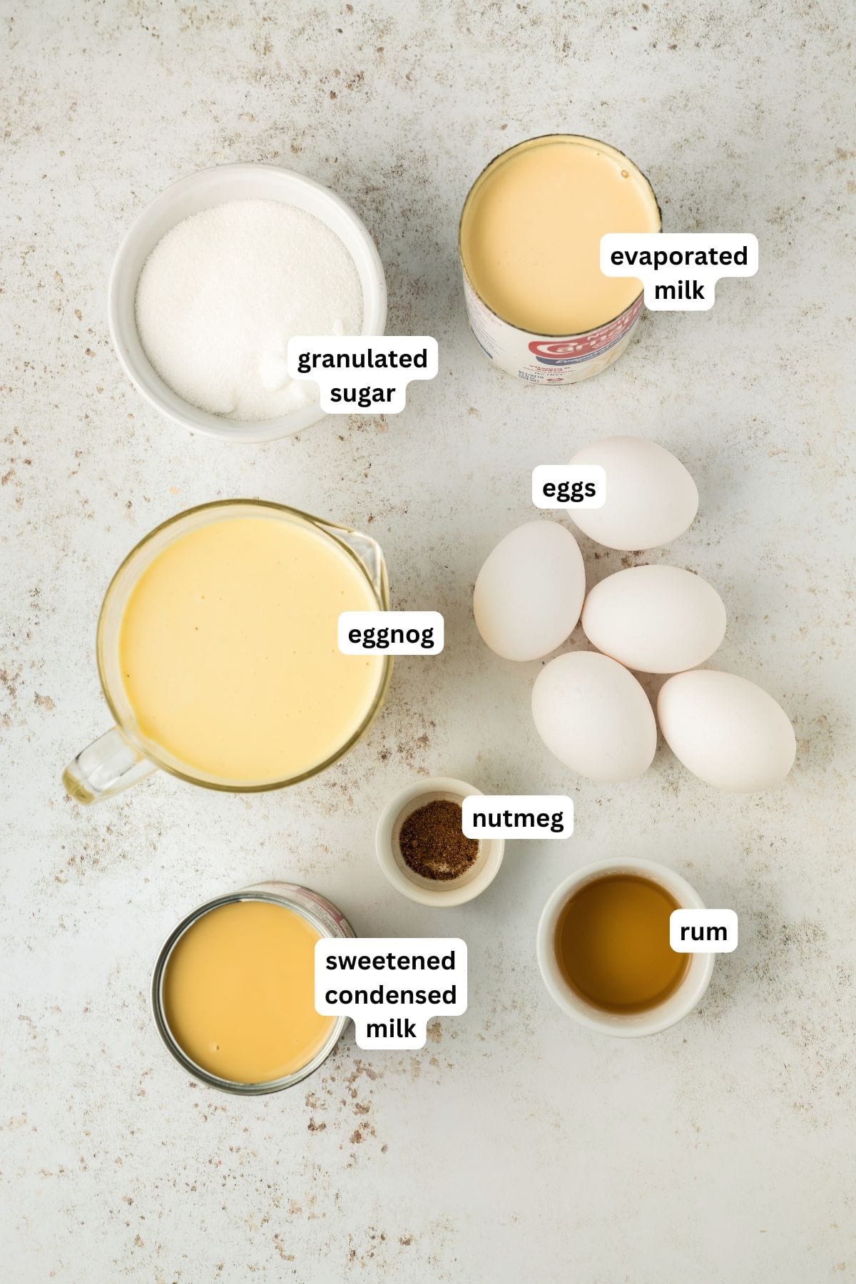 This festive Eggnog Flan is smooth, rich, and creamy, with plenty of nutmeg and rum for that classic spiced holiday flavor. Labeled ingredients for eggnog flan recipe are arranged on a light surface: granulated sugar, evaporated milk, eggs, eggnog, nutmeg, sweetened condensed milk, and rum (or rum extract).