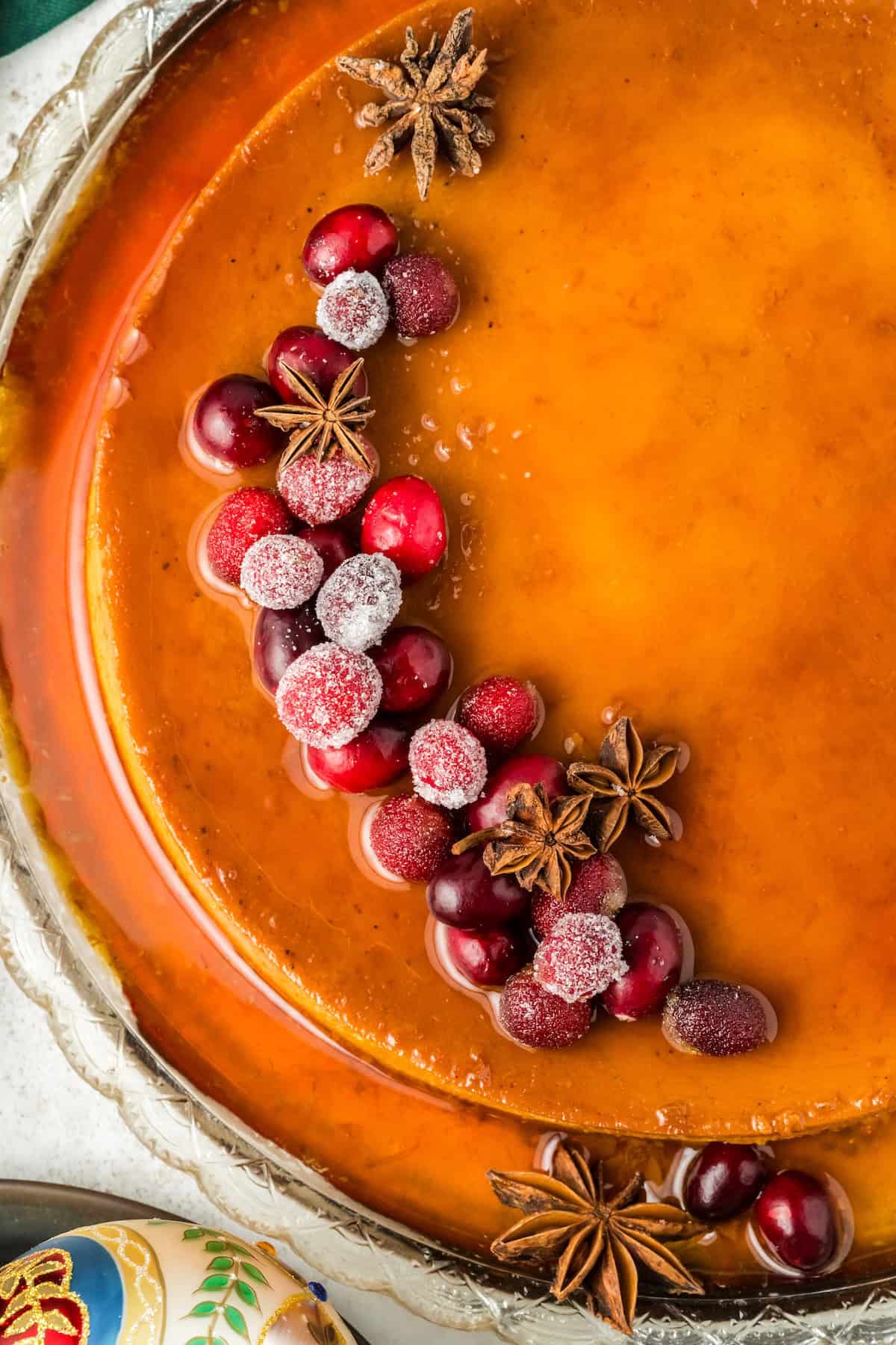 This festive Eggnog Flan is smooth, rich, and creamy, with plenty of nutmeg and rum for that classic spiced holiday flavor. A caramel colored eggnog flan topped with sugared cranberries and star anise, arranged decoratively on one side, served on a glass platter.