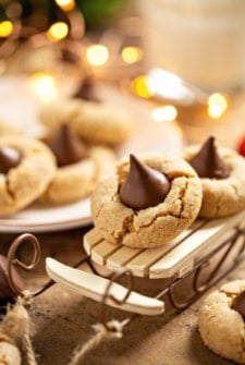 A close-up of soft peanut butter blossom cookies topped with chocolate kisses, one displayed on a small wooden sled.