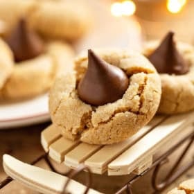 A peanut butter kiss cookie with a chocolate kiss on top sits on a small wooden sled, surrounded by more cookies.
