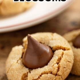 Close-up of a peanut butter blossom cookie with a chocolate kiss in the center, set on a wooden surface with another cookie and a plate in the background.
