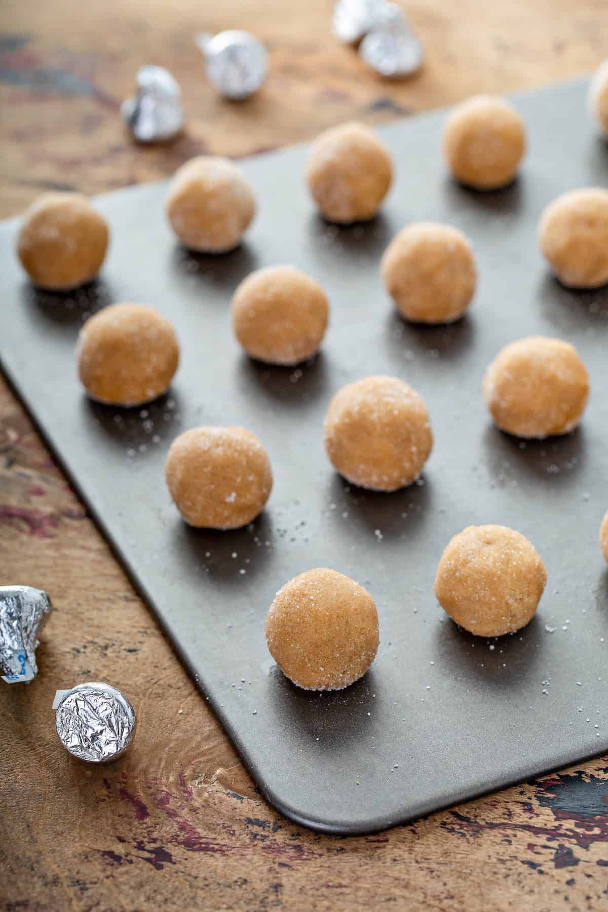These are the softest Peanut Butter Blossoms you’ll ever make! Perfect for holiday trays, neighbor gifts, or sneaking fresh from the oven. A baking sheet holds rows of unbaked peanut butter cookie dough balls dusted with sugar. Hershey’s Kisses candies wrapped in silver foil are scattered around.