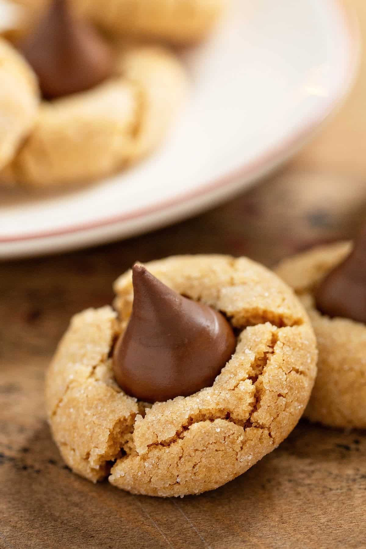 These are the softest Peanut Butter Blossoms you’ll ever make! Perfect for holiday trays, neighbor gifts, or sneaking fresh from the oven. A close-up of soft peanut butter blossom cookies topped with a chocolate kiss, with more cookies on a white plate in the background, set on a wooden surface.