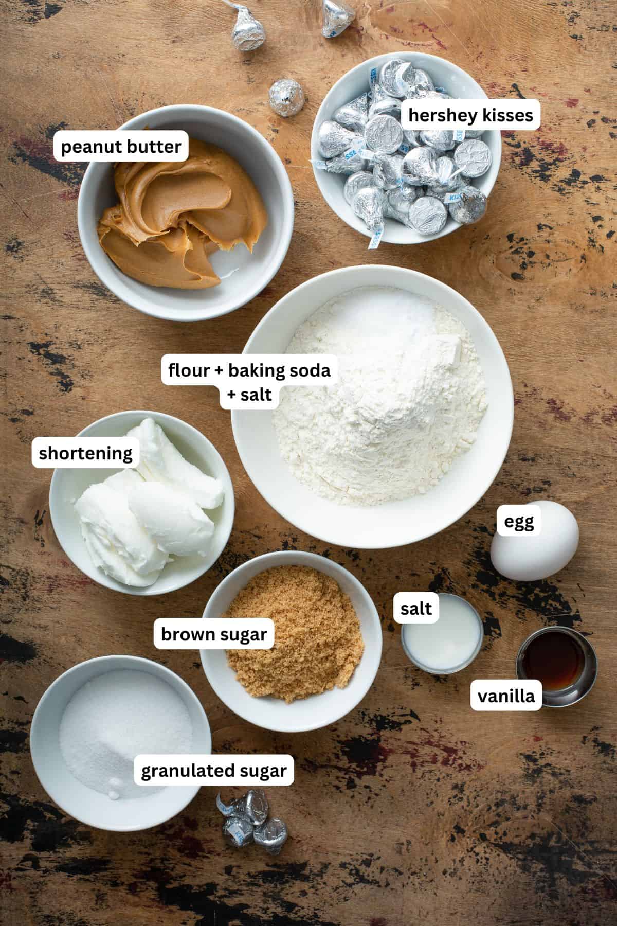 These are the softest Peanut Butter Blossoms you’ll ever make! Perfect for holiday trays, neighbor gifts, or sneaking fresh from the oven. Bowls containing labeled ingredients for this peanut butter blossoms recipe: peanut butter, Hershey Kisses, flour with baking soda and salt, shortening, brown sugar, granulated sugar, an egg, salt, and vanilla.