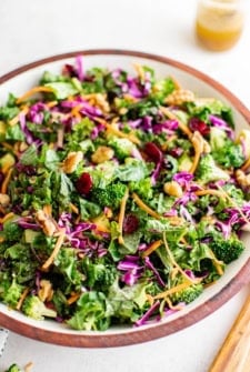 A vibrant chopped kale salad with cranberries, shredded red cabbage, carrots, broccoli, walnuts, and avocado in a large bowl with a jar of dressing in the background.