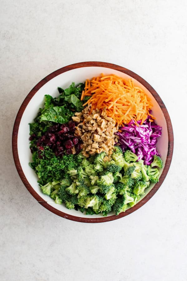 Broccoli Kale Salad with Cranberries is bright and colorful with a bold crunch and sweet pops of flavor. This salad always gets compliments! A large bowl filled with chopped kale, shredded carrots, chopped walnuts, dried cranberries, shredded purple cabbage, and broccoli florets, arranged in separate sections on a light surface.