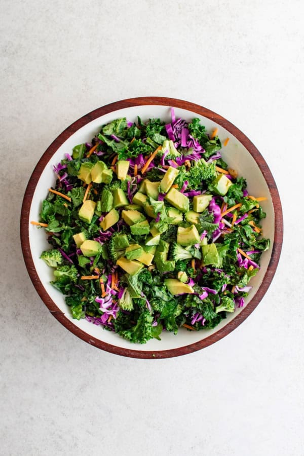 Broccoli Kale Salad with Cranberries is bright and colorful with a bold crunch and sweet pops of flavor. This salad always gets compliments! A wooden bowl filled with a colorful broccoli kale salad with cranberries, shredded purple cabbage, shredded carrots, and diced avocado on top, set on a light surface.