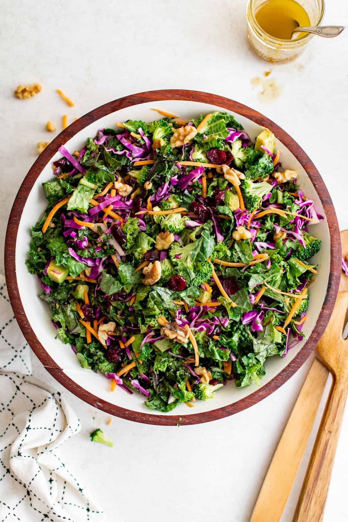Broccoli Kale Salad with Cranberries is bright and colorful with a bold crunch and sweet pops of flavor. This salad always gets compliments! A wooden bowl filled with broccoli kale salad, mixed with shredded carrots, red cabbage, walnuts, avocado, and dried cranberries. A small jar of dressing and wooden salad servers are set nearby on the side.