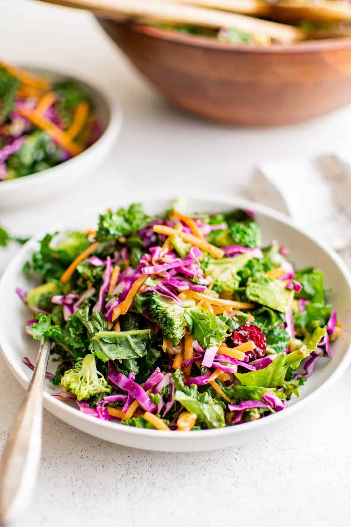 Broccoli Kale Salad with Cranberries is bright and colorful with a bold crunch and sweet pops of flavor. This salad always gets compliments! A bowl of colorful chopped kale salad with broccoli, shredded carrots, purple cabbage, avocado, and dried cranberries, served with a fork. Another bowl of salad and a wooden salad bowl are in the background.