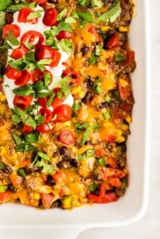 A close-up of a taco Dorito casserole topped with melted cheddar cheese, black beans, diced tomatoes, green onions, chopped cilantro, sliced cherry tomatoes, and a dollop of sour cream.