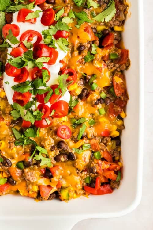 A close-up of a taco Dorito casserole topped with melted cheddar cheese, black beans, diced tomatoes, green onions, chopped cilantro, sliced cherry tomatoes, and a dollop of sour cream.