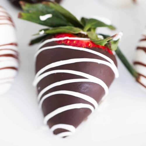 A fresh strawberry dipped in dark chocolate and drizzled with white chocolate, resting on a white surface with more chocolate-dipped strawberries in the background.
