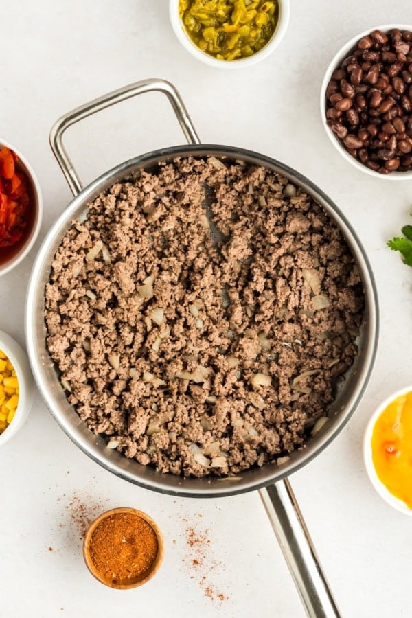 Crunchy, cheesy, beefy, and loaded with bold taco flavors, this Dorito Casserole recipe is quick to assemble and downright addictive! A skillet of cooked ground beef with onions is surrounded by small bowls of black beans, corn, diced tomatoes, chopped green chiles, shredded cheese, and taco seasoning on a white surface.