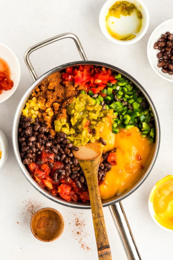 Crunchy, cheesy, beefy, and loaded with bold taco flavors, this Dorito Casserole recipe is quick to assemble and downright addictive! A pot filled with cooked ground beef, black beans, diced tomatoes, chopped red and green bell peppers, green chilies, yellow cheese sauce, and spices, with a wooden spoon for mixing. Small bowls of ingredients surround the pot.