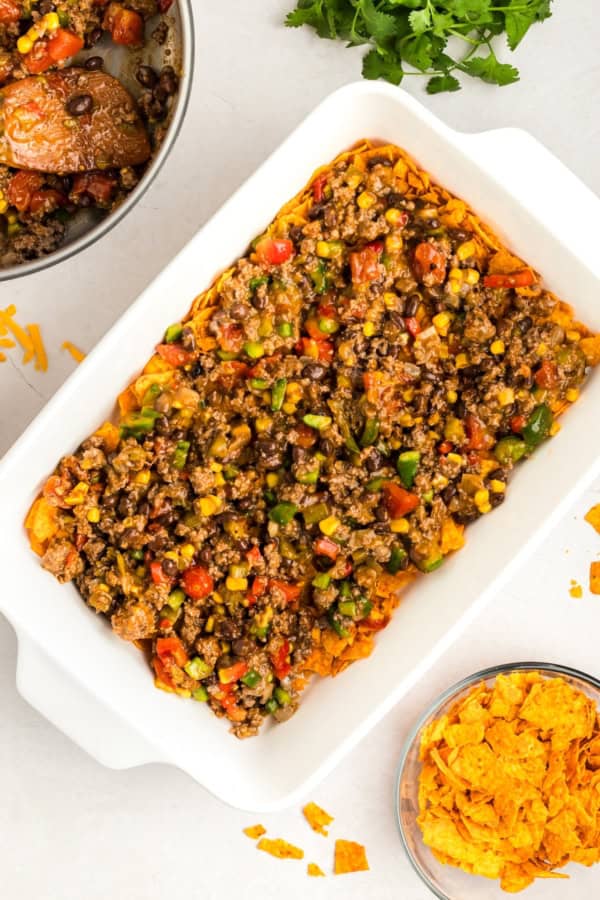 Crunchy, cheesy, beefy, and loaded with bold taco flavors, this Dorito Casserole recipe is quick to assemble and downright addictive! A white baking dish filled with crushed Doritos topped with a mixture of ground meat, black beans, corn, tomatoes, and green bell peppers.