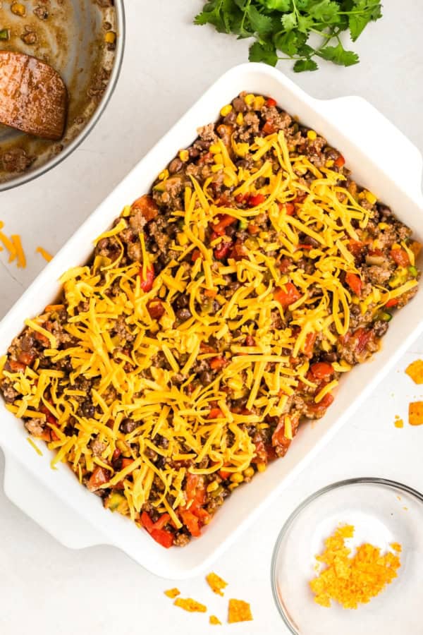 Crunchy, cheesy, beefy, and loaded with bold taco flavors, this Dorito Casserole recipe is quick to assemble and downright addictive! A white baking dish filled with a taco casserole with doritos, a layer of ground beef and vegetables mixture, and topped with shredded cheddar cheese.