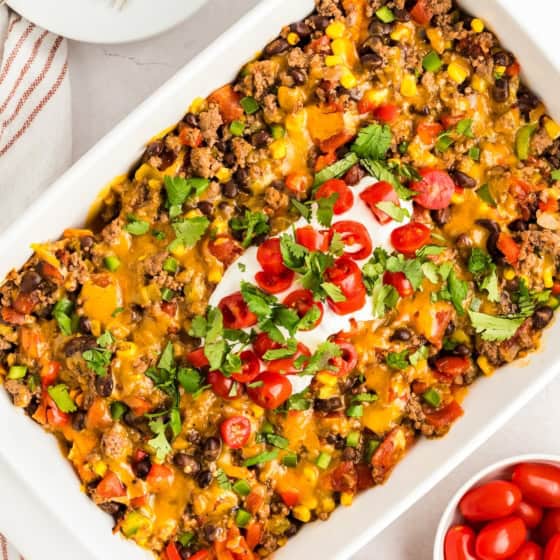 A baked taco casserole with Doritos in a white dish, topped with melted cheese, chopped tomatoes, cilantro, and a dollop of sour cream.